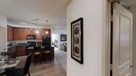 This single-floor residence showcases an open-concept layout that seamlessly connects the kitchen, living area, and bedroom. The kitchen is outfitted with rich, warm-toned wood cabinetry, black appliances including a refrigerator, stove, and microwave, and a breakfast bar with bar seating, making it both functional and inviting. Pendant lighting hangs above the bar area, complementing the recessed and track lighting overhead. The flooring transitions from wood-look planks in the kitchen and entryway to plush carpet in the living and bedroom areas, creating a comfortable and cohesive flow throughout. An arched doorway frames the transition between spaces, adding an architectural detail that enhances the home's character. The bedroom, visible through the open layout, appears well-proportioned and features neutral tones. Throughout the space, the walls are painted in a light, neutral palette, contributing to a bright and airy atmosphere. Decorative framed artwork on the wall near the entryway adds a finishing touch, reflecting a thoughtfully curated interior that balances practicality with aesthetic appeal.