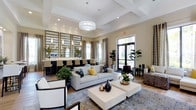 This single-floor space presents a polished and thoughtfully designed interior that blends comfort with contemporary elegance. The main lounge area is anchored by a coffered ceiling with recessed lighting and a statement pendant light fixture, lending a sense of architectural refinement and generous vertical volume. Warm neutral tones coat the walls, complemented by light hardwood-style flooring that extends throughout the space.

The seating area features a light gray sofa accented with yellow and patterned throw pillows, paired with white square coffee tables and additional accent chairs arranged around a decorative area rug. Tall indoor plants are strategically placed to introduce natural elements and enhance the inviting atmosphere.

Adjacent to the lounge, a bar-style counter area is lined with high-backed chairs and framed by a wall of mirrored panels hung with pendant lighting, creating a lively social zone. Large windows and glass doors along the perimeter flood the interior with natural light while offering views of the outdoors. Draped curtains frame the windows, adding a layer of texture and warmth to the overall design.