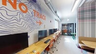 This dynamic single-floor workspace is designed with creativity and productivity in mind, combining energetic aesthetics with functional work areas. The main workstation zone features a long, natural wood benchtop lined with computer workstations and ergonomic orange rolling chairs, running alongside a white brick wall adorned with a bold "Leave No Trace" topographic mural rendered in navy blue and orange. Exposed silver ductwork runs overhead, contributing to the industrial character of the space, while pendant and recessed lighting ensure a well-lit environment throughout.

A glass-walled private meeting room provides a semi-enclosed collaborative option, furnished with orange chairs and a round table, offering visibility without sacrificing privacy. Adjacent to the main workstation run, a compact nook features built-in bench seating accented by a vibrant plaid-patterned partition, providing an informal breakout area.

The conference area is a standout feature, centered around a dramatic live-edge green epoxy resin table surrounded by tan leather chairs. Behind it, a large geometric owl mural on a wood-paneled accent wall adds artistic flair. A secondary workstation area benefits from wide windows with outdoor views and natural light, completing this visually rich and highly functional workspace.