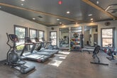 Located at 105 Oak Hill Dr in Athens, Georgia, this single-floor fitness center is thoughtfully designed to support a comprehensive workout experience. The space features rich, dark hardwood-style flooring throughout, complemented by a contrasting dark ceiling accented with exposed wooden beams and recessed lighting that creates a modern, focused atmosphere.

The main exercise area is well-equipped with multiple treadmills, an elliptical machine, and cycling bikes for cardiovascular training. A dedicated strength and functional training section includes a multi-station cable machine, a punching bag, and a rack stocked with kettlebells, medicine balls, and other free weights. Floor-to-ceiling mirrors along one wall visually expand the space and assist with form during workouts.

Natural light streams in through multiple large windows, offering views of the surrounding greenery and keeping the environment bright and inviting. A wall-mounted flat-screen television provides entertainment for cardio sessions.

Practical amenities include a double-spout water fountain conveniently positioned near the entrance, posted fitness center rules for user guidance, and clearly marked emergency exits. The overall design balances functionality with a clean, contemporary aesthetic, making this fitness center a well-rounded facility for residents and users of the Athens, GA community.