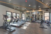 Located at 105 Oak Hill Dr in Athens, Georgia, this single-floor fitness center is thoughtfully designed to support a comprehensive workout experience. The space features rich, dark hardwood-style flooring throughout, complemented by a contrasting dark ceiling accented with exposed wooden beams and recessed lighting that creates a modern, focused atmosphere.

The main exercise area is well-equipped with multiple treadmills, an elliptical machine, and cycling bikes for cardiovascular training. A dedicated strength and functional training section includes a multi-station cable machine, a punching bag, and a rack stocked with kettlebells, medicine balls, and other free weights. Floor-to-ceiling mirrors along one wall visually expand the space and assist with form during workouts.

Natural light streams in through multiple large windows, offering views of the surrounding greenery and keeping the environment bright and inviting. A wall-mounted flat-screen television provides entertainment for cardio sessions.

Practical amenities include a double-spout water fountain conveniently positioned near the entrance, posted fitness center rules for user guidance, and clearly marked emergency exits. The overall design balances functionality with a clean, contemporary aesthetic, making this fitness center a well-rounded facility for residents and users of the Athens, GA community.