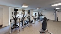 This comprehensive fitness center is thoughtfully designed to accommodate a wide range of workout styles and fitness goals. The facility is organized into distinct zones, each purpose-built and fully equipped. The cardio area features a row of elliptical trainers and treadmills positioned along windowed walls, allowing natural light to fill the space and providing an open, energizing atmosphere. Vertical blinds on the windows offer adjustable light control throughout the day.

A dedicated cycling studio is separated from the main floor by full-height glass partitions, creating a sleek, modern enclosure while maintaining visual connectivity. Inside, multiple stationary spin bikes are arranged facing a large wall-mounted display screen, beneath which a striking panoramic landscape mural adds an immersive quality to indoor cycling sessions.

The strength and functional training area hosts a multi-station cable machine, free weights on a tiered dumbbell rack, a weight bench, and an assortment of medicine balls, stability balls, and fitness accessories displayed on organized racks. The flooring throughout transitions between durable wood-look planks and carpet-tile zones, delineating different training areas.

Recessed panel lighting provides consistent, even illumination across all sections. The overall design is clean, bright, and professional, reflecting a facility built for serious, varied fitness use.