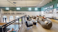 Located at 201 S 12th St in Bessemer City, NC, this thoughtfully reimagined industrial space blends historic character with contemporary design. The open-plan layout features soaring ceilings with exposed white-painted beams and visible ductwork, evoking its manufacturing heritage — further emphasized by a bold "Manufacturing Co." mural painted along the upper wall. Polished concrete floors anchor the space, while two-tone walls pair crisp white-painted brick below with a deep teal upper half, creating a striking visual contrast throughout. Natural light pours in through a row of clerestory windows positioned along the upper walls, complemented by sleek linear pendant lighting suspended from the ceiling. The lounge is furnished with a curated mix of modern seating, including curved bouclé sofas, leather-and-gold accent chairs, and round marble-top coffee tables. A built-in wooden bench with cushions and a marble-surface work table lines one wall, above which an extensive gallery wall displays framed historical photographs and documents. An open-railing mezzanine overlooking the level below adds verticality and depth. A dedicated seating nook with an orange accent chair adds a pop of color. The space offers a warm, creative atmosphere that honors its industrial past while delivering refined, contemporary comfort.