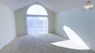 This single-floor space makes a striking architectural impression through its vaulted ceilings, which rise to a sharp peak and lend the interior a sense of openness and grandeur. The standout feature is a large arched window positioned at the apex of the vaulted wall, paired with full-height sliding glass doors beneath it. Together, these elements create a sweeping wall of natural light that bathes the room throughout the day. Vertical blinds provide adjustable light control while maintaining the airy feel of the space. The interior walls are finished in a clean, neutral white that reflects light and enhances the bright, open atmosphere. Soft, wall-to-wall carpeting in a warm beige tone covers the floors, adding a comfortable and understated quality to the space. The overall ambiance is calm and minimalist, with the architectural design doing much of the work. The combination of the vaulted ceiling, generous glazing, and neutral palette creates a versatile space well-suited to a variety of uses, offering both visual drama and a welcoming, light-filled environment.