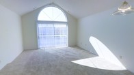 This single-floor space makes a striking architectural impression through its vaulted ceilings, which rise to a sharp peak and lend the interior a sense of openness and grandeur. The standout feature is a large arched window positioned at the apex of the vaulted wall, paired with full-height sliding glass doors beneath it. Together, these elements create a sweeping wall of natural light that bathes the room throughout the day. Vertical blinds provide adjustable light control while maintaining the airy feel of the space. The interior walls are finished in a clean, neutral white that reflects light and enhances the bright, open atmosphere. Soft, wall-to-wall carpeting in a warm beige tone covers the floors, adding a comfortable and understated quality to the space. The overall ambiance is calm and minimalist, with the architectural design doing much of the work. The combination of the vaulted ceiling, generous glazing, and neutral palette creates a versatile space well-suited to a variety of uses, offering both visual drama and a welcoming, light-filled environment.