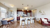 Located at 100 Seasons Blvd in Dundee, IL, this single-floor residence offers a thoughtfully designed open-concept layout with contemporary finishes throughout. The kitchen serves as a central hub, showcasing rich dark-wood cabinetry, white quartz countertops, stainless steel appliances including a range, microwave, and refrigerator, and a large center island with pendant lighting and bar seating. The kitchen flows seamlessly into a dining area and a bright living room, where large windows with plantation shutters invite abundant natural light and views of the surrounding neighborhood. Wood-look flooring runs throughout the main living areas, adding warmth and continuity to the space. The primary bedroom is generously sized, featuring plush carpeting, oversized windows, and access to a well-appointed en-suite bathroom with a dark-wood vanity, white countertop, and a walk-in closet with built-in shelving. A second bedroom offers comfortable proportions with its own window and carpeted floors. A second full bathroom includes a tub-shower combination and matching vanity finishes. An in-unit laundry area adds everyday convenience. The overall design balances modern aesthetics with a warm, livable ambiance throughout.
