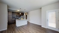 This single-floor residence offers a thoughtfully designed open-concept layout that seamlessly connects the kitchen and living areas, creating a bright and airy atmosphere. The kitchen is a standout feature, equipped with stainless steel appliances including a refrigerator, microwave, and range. Rich dark cabinetry provides ample storage and contrasts elegantly with the light quartz countertops. A central kitchen island serves as both a functional workspace and a casual gathering spot, accented by stylish pendant lighting that hangs overhead. Additional flush-mount ceiling lighting ensures the space remains well-lit throughout. The living area extends naturally from the kitchen, offering generous open space defined by smooth, neutral-toned walls and clean white baseboards. Warm hardwood-style plank flooring runs continuously across the entire space, adding texture and cohesion to the overall design. The gently curved wall transition between the kitchen and living areas adds an architectural softness to the layout. The overall ambiance is modern, clean, and inviting, with a neutral color palette that provides a versatile backdrop suitable for a variety of interior styles and furnishings.