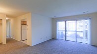 This single-story residence features an open-concept layout that connects the living area to the kitchen, creating a functional and cohesive living space. The carpeted living room is generously sized, finished with neutral white walls and a smooth ceiling that contribute to a clean, understated aesthetic. A full-width sliding glass door lined with vertical blinds serves as both a prominent source of natural light and a direct access point to an outdoor area, where a patio or balcony is visible beyond. The abundance of natural light lends the space a bright, airy quality.

Adjacent to the living area, the kitchen is partially visible through an open passageway, revealing warm wood-toned cabinetry and a white refrigerator. The layout allows for easy flow between the kitchen and living spaces, making the unit practical for everyday use. The overall design is straightforward and well-maintained, with simple finishes throughout. The single-floor layout ensures ease of movement, while the connection to an outdoor space adds a desirable element of openness to the unit's overall feel.