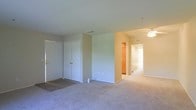 This single-floor residence offers a straightforward, functional layout designed for comfortable everyday living. The main living area is spacious and open, featuring wall-to-wall neutral carpeting that lends a warm, cohesive feel throughout the space. A set of built-in closet doors near the entryway provides convenient storage directly off the main living area, adding to the unit's practicality.

The interior is finished with light, neutral-toned walls that reflect natural light and create a clean, airy atmosphere. A ceiling fan is mounted overhead, offering both lighting and airflow for year-round comfort. Visible HVAC vents integrated into the walls and ceiling indicate a centralized climate control system.

A hallway extends from the main living space, leading to additional rooms at the rear of the residence. Glimpses through the hallway reveal what appears to be a closet or storage nook with warm wood-toned finishes, along with access to further living spaces. The layout flows in a logical and connected manner, making the space feel cohesive and easy to navigate.

Overall, the residence presents a clean, move-in ready environment with a neutral palette, practical storage solutions, and a well-organized floor plan suited for a range of occupants.