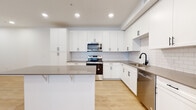 This single-floor residence showcases a clean, contemporary design with a consistent neutral palette and modern finishes throughout. The kitchen serves as a central highlight, featuring an abundance of white shaker-style cabinetry extending to the ceiling for maximum storage. Stainless steel appliances, including a range, microwave, dishwasher, and refrigerator, are seamlessly integrated alongside a white subway tile backsplash and grey quartz countertops. A substantial island provides additional prep and seating space. Light wood-tone flooring runs throughout the main living and kitchen areas, enhancing the bright, open atmosphere created by recessed lighting.

The bedrooms offer plush carpeting, natural light through slatted blinds, and generous closet space with built-in shelving and hanging rods. The primary bathroom is notably spacious, featuring a glass-enclosed shower, a separate soaking tub, a white vanity with grey countertop, and direct access to a walk-in closet. A secondary bathroom mirrors the same design aesthetic, with a white shaker vanity, grey countertop, black hardware accents, and a tub-shower combination. Together, the spaces present a cohesive, well-finished environment suited for modern living.