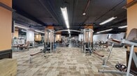 Located at 200 N Harvey Ave in downtown Oklahoma City, this fitness facility offers a generously sized, single-floor workout space designed to accommodate a full spectrum of strength and conditioning training. The open floor plan is anchored by distinctive orange-accented structural columns that provide visual definition and divide the space into functional training zones without interrupting the overall flow.

The facility is outfitted with an extensive array of professional-grade equipment, including cable machines, multi-station cable crossover units, selectorized resistance machines, free-weight benches, squat racks, and a well-stocked dumbbell and kettlebell section. Flooring transitions strategically between patterned carpet tile in the machine areas and durable rubber flooring in the free-weight zones, optimizing both comfort and safety.

Overhead, an exposed dark ceiling houses industrial-style linear LED lighting that illuminates the entire floor evenly. Wall-mounted sconces on the orange columns add a warmer ambient tone. Full-length mirrors are positioned throughout, supporting proper form and enhancing the sense of depth and spaciousness. Wall-mounted monitors are also present for entertainment or informational use. The overall ambiance is functional and professional, suited for serious athletic training in an urban setting.