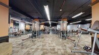 Located at 200 N Harvey Ave in downtown Oklahoma City, this fitness facility offers a generously sized, single-floor workout space designed to accommodate a full spectrum of strength and conditioning training. The open floor plan is anchored by distinctive orange-accented structural columns that provide visual definition and divide the space into functional training zones without interrupting the overall flow.

The facility is outfitted with an extensive array of professional-grade equipment, including cable machines, multi-station cable crossover units, selectorized resistance machines, free-weight benches, squat racks, and a well-stocked dumbbell and kettlebell section. Flooring transitions strategically between patterned carpet tile in the machine areas and durable rubber flooring in the free-weight zones, optimizing both comfort and safety.

Overhead, an exposed dark ceiling houses industrial-style linear LED lighting that illuminates the entire floor evenly. Wall-mounted sconces on the orange columns add a warmer ambient tone. Full-length mirrors are positioned throughout, supporting proper form and enhancing the sense of depth and spaciousness. Wall-mounted monitors are also present for entertainment or informational use. The overall ambiance is functional and professional, suited for serious athletic training in an urban setting.