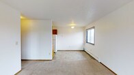 This single-floor residential space presents a straightforward and functional layout designed for comfortable everyday living. The main living area is finished with wall-to-wall carpeting, contributing a warm and understated feel to the room. A flush-mounted ceiling light provides overhead illumination, while a sliding window along one wall allows natural light to filter in and provides ventilation. A baseboard heating unit runs along the exterior wall, ensuring year-round comfort. The textured ceiling adds a subtle layer of detail to the otherwise clean, white-painted walls. Transitioning from the living area, the flooring shifts to a tile pattern, delineating the kitchen space. The kitchen is equipped with essential appliances, including a full-size refrigerator, and features rich, dark-toned cabinetry mounted above the workspace, offering practical storage. The open sightline between the living and kitchen areas creates a sense of connected, efficient use of space. Overall, the unit presents a clean, no-frills environment well-suited for those seeking a practical and easy-to-maintain living space.