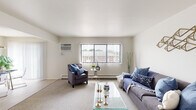 This single-floor residence offers a well-organized layout designed for comfortable everyday living. The living room serves as the central gathering space, featuring plush carpeting, a large window with vertical blinds that invites natural light and outdoor views, and a wall-mounted air conditioning unit alongside baseboard heating. The open, neutral-toned walls create a versatile backdrop adaptable to various décor styles.

Adjacent to the living room, the dining area connects seamlessly to the kitchen via an open pass-through, enhancing the sense of flow. A pendant light anchors the dining space, which benefits from additional natural light through a sliding panel window. The kitchen is equipped with dark cabinetry, light countertops, a full-size refrigerator, electric range, and a stainless steel sink — all set on durable wood-look flooring.

The two bedrooms are carpeted and each includes a window and baseboard heater. One bedroom is generously proportioned enough to accommodate multiple beds. The bathroom features warm tile flooring, dark vanity cabinetry, and a clean, functional layout. Throughout the unit, a consistent neutral color palette and practical finishes contribute to a bright, cohesive ambiance.