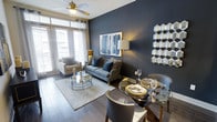 This single-floor space showcases a thoughtfully designed open-concept layout that seamlessly integrates the living and dining areas. The interior is defined by rich hardwood flooring that runs throughout, lending warmth and continuity to the space.

The living area is anchored by a bold navy accent wall, which serves as a striking backdrop for a contemporary blue sofa adorned with coordinating throw pillows. A plush area rug with a geometric pattern grounds the seating arrangement, complemented by a circular glass coffee table. A ceiling fan with a brushed gold finish adds both function and style overhead, while large windows dressed in sheer curtains allow generous natural light to flood the room. A cozy armchair positioned near the windows creates an ideal reading nook.

The dining area features a round glass-top table paired with sleek upholstered chairs, elegantly set with gold-accented dinnerware. A geometric honeycomb mirror with gold and dark tones adorns the wall, reinforcing the space's sophisticated aesthetic. Together, the navy, gold, and neutral tones create a cohesive, upscale ambiance throughout.