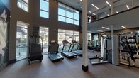 Located at 305 W Commerce St in Dallas, TX, this two-story fitness center is a well-appointed facility designed to support a comprehensive range of workout routines. The main level is anchored by a cardio zone featuring multiple treadmills, elliptical trainers, and rowing machines, all positioned along expansive floor-to-ceiling windows that flood the space with natural light and offer views of the surrounding landscaped outdoor areas. A dedicated strength training section is equipped with cable machines, free weights, and functional training equipment, complemented by full-length mirrors and linear LED lighting overhead. A separate studio space on this level accommodates spin bikes and additional cardio equipment, with a large display screen and mirrored walls enhancing the workout environment. A modern staircase with sleek cable railings connects the two floors, leading to an upper mezzanine level that provides additional equipment and overlooks the main floor below. The upper level is distinguished by a bold, large-scale graphic mural rendered in monochromatic tones, adding a striking artistic element to the space. Throughout, the facility maintains a contemporary aesthetic with neutral tones, durable flooring, and recessed and linear lighting, creating a functional yet visually engaging environment for fitness.