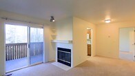 This single-floor space is designed with a practical and open layout that maximizes comfort and functionality. The living area serves as the heart of the unit, centered around a fireplace framed by a clean white mantel and tiled hearth, providing a classic focal point to the room. Above the fireplace, a built-in shelf adds a subtle decorative element. The living space is finished with neutral-toned carpet, contributing to a warm and understated aesthetic throughout.

Natural light flows generously into the room through a large sliding glass door fitted with sheer curtains, which opens to an outdoor balcony with a slatted railing, offering views of mature trees beyond. A track light fixture mounted on the ceiling provides additional illumination when needed.

Adjacent to the living area, a passthrough opening leads into the kitchen, which features warm wood-toned cabinetry and white appliances, including what appears to be a refrigerator and microwave. The kitchen is compact yet well-organized, with cabinetry extending to the upper walls for ample storage. The overall palette of the space — soft whites, beige tones, and warm wood accents — creates a cohesive and inviting environment.