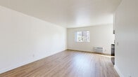 This single-floor unit presents a clean, minimalist aesthetic defined by crisp white walls, smooth ceilings, and warm light hardwood flooring that runs consistently throughout the space. The open living area is generously proportioned, with natural light streaming in through multiple windows that offer views of the surrounding neighborhood. A wall-mounted HVAC unit provides climate control directly within the main living area.

The kitchen is thoughtfully designed and well-equipped, featuring white shaker-style cabinetry with sleek hardware, a stainless steel sink with a gooseneck faucet, a stainless steel gas range with oven, and an overhead range hood. A window allows natural light to fill the kitchen, enhancing the bright and airy atmosphere. The layout offers a practical workflow, with ample counter and cabinet space for everyday use.

A separate bedroom provides a quiet retreat, continuing the same light hardwood flooring and neutral white walls, creating a calm and versatile space. The bathroom is clean and functional, equipped with a full-size soaking tub and shower combination with chrome fixtures and a crisp white surround. Throughout the unit, the consistent use of light tones and natural materials creates a cohesive, welcoming ambiance suited for comfortable everyday living.