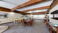 Located on Paramount Blvd in Downey, California, this single-floor community space offers a well-organized layout designed for gathering and recreation. The space is defined by its striking exposed wood beam ceilings, which lend a warm, rustic character throughout and create a sense of architectural depth.

The expansive game room serves as the heart of the space, accommodating a ping-pong table, a pool table, and multiple dining and seating areas. A central pendant-lit suspended fireplace serves as a focal point, adding ambiance to the room. Neutral-toned carpet covers the floor, while white-paneled walls and large windows with vertical blinds allow for ample natural light. A wall-mounted television and additional seating arrangements make this room well-suited for both active recreation and casual socializing.

The kitchen is efficiently designed with a U-shaped layout featuring white cabinetry, a double stainless steel sink, a coil-top electric range, a refrigerator, and an over-range ventilation hood. A pass-through counter connects the kitchen to the adjacent dining area, facilitating easy service during events. Wall sconce lighting and the continuation of the exposed beam ceiling maintain visual consistency throughout the space. Overall, the layout is practical and functional, making it an ideal venue for community events and group activities.