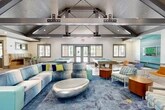 Located at 1001 Southwest 17th Lane in Gainesville, Florida, this single-floor community clubhouse offers a generously sized, open-concept gathering space designed for socializing and recreation. The architectural centerpiece is a dramatic vaulted ceiling with exposed steel truss work and a wood-paneled interior, creating an airy, loft-like atmosphere that fills the space with a sense of grandeur. Large gray-trimmed windows and glass double doors line the walls, flooding the interior with natural light and providing views of the outdoor pool area beyond. The main lounge area features a large sectional sofa accented with colorful throw pillows, arranged around a distinctive silver oval coffee table atop a bold blue area rug. Multiple billiard tables with bright blue felt are positioned throughout, adding a recreational focal point to the space. Light wood-look tile flooring extends across much of the floor, complemented by a mix of modern seating including modular chairs in vibrant orange and blue tones. A dedicated media nook showcases a large flat-screen TV mounted on a striking turquoise shiplap accent wall. Semi-private alcoves with decorative slatted partitions and bold painted murals provide additional nooks for smaller gatherings. A built-in banquette seating area and a bar-height counter with colorful stools complete the amenity-rich layout.