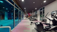 Located at 10920 Garfield Ave in South Gate, Los Angeles, this single-floor fitness center is a comprehensively outfitted facility designed to support a wide range of workout routines. The space is defined by a contemporary aesthetic, featuring dark ceilings with recessed lighting that creates a focused, energetic atmosphere throughout. A distinctive, colorful mural spanning one wall serves as a bold visual centerpiece, adding a dynamic artistic element to the environment.

The cardio section is generously appointed with multiple treadmills, elliptical trainers, stationary bikes, rowing machines, and StairMaster units arranged in well-organized rows. Floor-to-ceiling glass windows and curtain wall panels line the perimeter, offering views of the exterior and allowing natural light to supplement the ambient recessed lighting.

A dedicated training zone, clearly marked and enclosed by glass partitions, houses a robust free weight area with a full rack of dumbbells, cable machines, a power rack, TRX suspension trainers, and functional training equipment including a BOSU ball and a Vasa trainer. The flooring throughout features a patterned pink and grey carpet tile design that delineates different workout zones while contributing to the facility's modern visual identity. The overall layout is spacious, organized, and well-suited for both cardio-focused and strength-based training.
