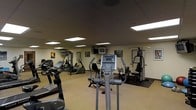 This single-floor fitness room offers a practical and functional environment designed to support a range of workout routines. The space is outfitted with a diverse selection of exercise equipment, including treadmills, elliptical trainers, a recumbent bike, and multi-station cable weight machines, providing options for both cardiovascular and strength training exercises. A large mirror runs along one wall, visually expanding the room and allowing users to monitor their form during workouts. The ceiling features a dark drop-tile finish with evenly spaced recessed panel lights that cast a bright, consistent glow throughout the space. Wall-mounted televisions are positioned for easy viewing during cardio sessions, adding an entertainment element to the workout experience. Framed artwork and informational posters decorate the walls, contributing a modest degree of visual interest. Rubber floor mats are placed beneath the heavier equipment to protect the flooring and reduce noise. The overall layout is organized and functional, making efficient use of the available floor space while maintaining clear pathways between equipment stations.