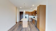 This single-floor unit showcases a clean, contemporary design with a cohesive aesthetic throughout. The open-concept kitchen serves as the heart of the space, outfitted with flat-front warm wood cabinetry that extends floor to ceiling, offering generous storage. A suite of stainless steel appliances includes a French door refrigerator with ice dispenser, an electric range with a stainless hood vent, a built-in microwave, and a dishwasher. A white quartz countertop island anchors the kitchen, illuminated above by two elegant glass pendant lights. The cabinetry also incorporates a built-in wine rack, adding a thoughtful design detail. Wood-look luxury vinyl plank flooring runs throughout the main living areas, complementing the warm tones of the cabinetry.

The living area extends toward a sliding glass door leading to a balcony, providing city views and natural light. The bedroom features plush carpeting, a window framing outdoor views, and a well-organized closet with sliding doors, multiple hanging rods, and built-in shelving. The bathrooms maintain the modern aesthetic with wood-toned vanities, white quartz countertops, backlit mirrors, subway-tiled tub surrounds, and wood-look flooring.