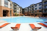 This expansive outdoor amenity space centers around a large, resort-style swimming pool with crystal-clear blue water, set within a beautifully designed courtyard. The surrounding multi-story building features a striking combination of warm brick masonry and modern light-colored paneling, with private balconies overlooking the pool deck on every level, creating an enclosed, resort-like atmosphere.

The generous pool deck is finished in light-toned concrete pavers, providing ample space for relaxation and socializing. Stylish chaise lounge chairs with bold orange cushions are arranged throughout the deck, offering both sun and shade options. A dedicated sun shelf area at the pool's edge allows for in-water lounging, while nearby pergola structures provide shaded retreats accented by orange market umbrellas.

A secondary lounge courtyard adjacent to the main pool area features additional seating arrangements with orange-cushioned chairs, low tables, and lush landscaping including ornamental shrubs and planted beds along the building's brick facade. Floor-to-ceiling windows and glass-paneled doors line the ground level, connecting interior amenity spaces directly to the outdoor area and filling both zones with natural light. The overall ambiance is modern, vibrant, and well-appointed, designed to encourage an active and social outdoor lifestyle.