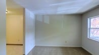 This single-floor space presents a straightforward and functional layout with a clean, move-in-ready aesthetic. The main room is finished with wall-to-wall carpet in a neutral tone, providing a comfortable underfoot feel while complementing the light-colored walls. The walls are painted in a soft, understated hue that lends a calm and versatile backdrop suitable for a variety of uses or décor styles.

The ceiling features recessed panel lighting, which provides even, ambient illumination throughout the room. The reflective quality of the ceiling finish helps distribute light effectively, enhancing the overall brightness of the interior. Baseboards run cleanly along the perimeter of the room, adding a polished, finished look to the space.

A partially visible adjacent area to the left suggests additional rooms or transitional spaces within the floor plan, hinting at a connected layout. The room includes a standard electrical outlet along the lower wall, reflecting practical utility considerations. Overall, the space offers a blank canvas with neutral finishes and adequate lighting, making it highly adaptable for residential or light commercial purposes.
