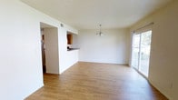 This two-floor space offers a functional and well-connected layout beginning with an open-plan living and dining area. The room is finished with light-toned hardwood-style flooring that runs throughout, contributing to a clean and cohesive aesthetic. A modest chandelier serves as a focal point above the dining area, while a large sliding glass door along one wall allows generous natural light to fill the space and provides access to an outdoor area.

The kitchen is positioned adjacent to the living and dining area and features a pass-through counter opening, allowing visual connectivity between the spaces. Warm-toned wood cabinetry lines the kitchen walls, complemented by a dark countertop. A stainless steel refrigerator is visible, and additional appliances and counter space are accessible within the kitchen footprint.

The ceilings feature a textured finish throughout the main level, and the walls are painted in a neutral white tone, providing a versatile backdrop. HVAC venting is integrated into the ceiling, ensuring climate control throughout the space. The overall ambiance is bright, open, and practical, suited for everyday living across two floors.