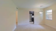 This single-floor space offers a clean, functional layout with an open feel enhanced by light-colored walls and neutral tile flooring. The main living area is bright and airy, with smooth white ceilings and crisp white baseboards that contrast neatly against the warm beige walls. A pendant light fixture with a bowl-shaped shade hangs overhead, providing a subtle focal point and ambient lighting to the space.

The kitchen is visible through an open passageway, creating a connected flow between the living and cooking areas. It features dark espresso-toned cabinetry that extends to the upper walls, offering generous storage. A stainless steel refrigerator anchors the space, complemented by a sink and dark countertops, giving the kitchen a contemporary and practical character. The kitchen flooring mirrors the light tile found throughout the rest of the unit, maintaining visual consistency.

A hallway extends off the main living area, suggesting access to additional rooms beyond what is visible. The overall ambiance of the space is modest yet well-maintained, with a neutral palette that provides a versatile backdrop suited to a variety of interior styles.