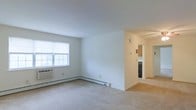 This single-floor space presents a clean, functional layout centered around an open living area finished with wall-to-wall neutral carpeting. The room is characterized by crisp white walls and a smooth ceiling, contributing to a bright and airy atmosphere. A window on the exterior wall allows natural light to filter into the space, offering a view of surrounding greenery. Climate control is provided by a wall-mounted air conditioning unit beneath the window, complemented by a baseboard heating system that runs along the perimeter of the room — practical features ensuring year-round comfort. The living area flows naturally toward an adjacent hallway, which leads to a kitchen area visible in the background. The kitchen features warm-toned wood cabinetry and dark appliances, offering a functional cooking space. The overall design is straightforward and utilitarian, with a neutral color palette throughout that lends itself well to personalization. The transition between the carpeted living space and what appears to be a hard-surface flooring in the kitchen and hallway area adds subtle definition between the functional zones of this single-level layout.