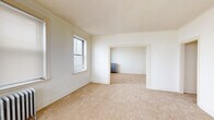 Located at 7447 S South Shore Dr in Chicago's South Shore neighborhood, this single-floor unit offers a well-defined layout with multiple interconnected rooms. The interior is characterized by neutral white walls, textured ceilings, and warm beige carpeting throughout the living and sleeping areas, creating a clean and functional atmosphere.

The living spaces are generously proportioned, with multiple windows fitted with blinds that allow for adjustable natural light. Classic cast-iron radiators are present throughout, providing consistent radiant heat and serving as a nod to the building's architectural heritage. Detailed door and window trim adds a traditional character to each room.

A central hallway connects the rooms and leads to the front entry door, providing a clear and practical flow through the unit. Wall-mounted light fixtures illuminate the corridors efficiently.

The kitchen is equipped with white cabinetry featuring wood trim accents, a stainless steel refrigerator, a stainless steel gas range, and a built-in shelving nook. Tile-pattern flooring and a window above the work area complete the functional kitchen space.

The bathroom features a classic soaking tub with a shower, white tile surround, a vanity with mirror, terrazzo-style flooring, a window for natural light, and a radiator for warmth. Additional closet and storage space is accessible throughout the unit.