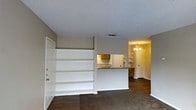 This single-floor residence in Tyler, Texas offers a thoughtfully arranged open-concept layout that connects the main living area to the kitchen and dining spaces. The living room features built-in white shelving along one wall, providing ample display and storage space, and is finished with neutral beige tones and dark wood-style flooring that adds warmth and contrast. A textured ceiling runs throughout, lending a classic residential character to the space. The kitchen is accessible via a pass-through counter with a dark countertop, allowing for easy interaction between the cooking and living areas. Wood-toned cabinetry and open shelving in the kitchen provide functional storage. Moving toward the dining area, an elegant chandelier serves as a focal point, casting a welcoming glow over the space. The flooring transitions into a warm carpet, complementing the neutral wall palette. The overall ambiance is cozy and functional, with a layout that promotes a natural flow from room to room. Built-in details and decorative lighting elevate the everyday practicality of this single-story home, making it a comfortable and well-appointed space in the heart of Tyler, TX.