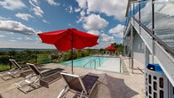 Located at 900 Luxor Ln in Norristown, PA, this expansive outdoor pool and terrace area offers a resort-style amenity space spanning an impressive footprint. The centerpiece of this outdoor retreat is a sleek infinity-edge pool with crystal-clear water, designed to visually merge with the sweeping panoramic views of the surrounding landscape, including rolling greenery and the broader Norristown skyline beyond.

The pool deck is finished with a neutral stone-toned surface and is generously appointed with modern sling-style lounge chairs and bold red market umbrellas, providing ample space for relaxation and sunbathing. Steel-framed staircases with glass panel railings connect the pool level to upper and lower terraces, adding a contemporary architectural edge to the overall design.

Adjacent to the pool, an open-air terrace features high-top dining tables and chairs, surrounded by young landscaped trees and framed by sleek metal railings that preserve the unobstructed views of the horizon. The elevated position of this outdoor space ensures that both the pool and terrace areas benefit from wide-open sightlines across the treetops and neighboring community.

The multi-story building backdrop, clad in a mix of tan and red exterior panels with large windows, anchors the space architecturally, while the overall design creates a vibrant and functional outdoor amenity ideal for both leisure and social gatherings.