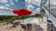Located at 900 Luxor Ln in Norristown, PA, this expansive outdoor pool and terrace area offers a resort-style amenity space spanning an impressive footprint. The centerpiece of this outdoor retreat is a sleek infinity-edge pool with crystal-clear water, designed to visually merge with the sweeping panoramic views of the surrounding landscape, including rolling greenery and the broader Norristown skyline beyond.

The pool deck is finished with a neutral stone-toned surface and is generously appointed with modern sling-style lounge chairs and bold red market umbrellas, providing ample space for relaxation and sunbathing. Steel-framed staircases with glass panel railings connect the pool level to upper and lower terraces, adding a contemporary architectural edge to the overall design.

Adjacent to the pool, an open-air terrace features high-top dining tables and chairs, surrounded by young landscaped trees and framed by sleek metal railings that preserve the unobstructed views of the horizon. The elevated position of this outdoor space ensures that both the pool and terrace areas benefit from wide-open sightlines across the treetops and neighboring community.

The multi-story building backdrop, clad in a mix of tan and red exterior panels with large windows, anchors the space architecturally, while the overall design creates a vibrant and functional outdoor amenity ideal for both leisure and social gatherings.