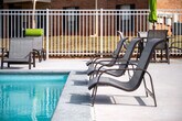 This outdoor pool area offers a well-appointed recreational space designed for relaxation and leisure. The centerpiece is a generously sized rectangular swimming pool with clear, turquoise water, accessible via stainless steel handrail entries on multiple sides. The pool's clean lines and bright water create a welcoming focal point for the entire area.

Surrounding the pool is a broad concrete sundeck that provides ample space for movement and lounging. Multiple cushioned chaise lounge chairs, accented with bright green cushions, are arranged along the poolside, offering comfortable spots for sunbathing. Additional seating options are distributed around the perimeter to accommodate various preferences.

A brick building with a pitched roof sits directly adjacent to the pool deck, likely serving as a clubhouse or amenity facility. Its large windows and glass doors open toward the pool area, reinforcing the connection between indoor and outdoor spaces.

The entire pool area is enclosed by a white metal fence, providing both security and a clean, defined boundary. Mature trees frame the backdrop, offering partial shade and a natural aesthetic. The surrounding residential brick buildings are visible beyond the fence, providing context within the broader community. Open skies above ensure abundant natural light throughout the day, enhancing the overall outdoor ambiance.