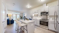 This single-floor residence offers a bright, modern layout designed for comfortable everyday living. The open-concept kitchen is a standout feature, outfitted with shaker-style cabinetry in a soft grey-white finish, stainless steel appliances including a refrigerator, range, microwave, and dishwasher, and a white quartz-topped island with bar seating. Light wood-look flooring runs throughout the kitchen and living areas, contributing to a clean, cohesive aesthetic.

The living room flows naturally from the kitchen and is bathed in natural light from a set of glass patio doors that open to an outdoor balcony. The space is generous enough to accommodate a full sofa arrangement and accent seating, with neutral walls providing a versatile backdrop.

The bedroom is a calm retreat featuring plush carpeting, a large window with views of the outdoors, and ample wall space. An adjacent nook or den area adds flexible living space, accented by light hardwood-style flooring.

The bathroom is designed with accessibility in mind, featuring grab bars near the shower and toilet, a wide-format vanity with grey cabinetry and a white countertop, and light vinyl flooring. Throughout the residence, the design maintains a cohesive palette of whites, soft greys, and warm neutrals, creating an inviting and functional living environment.