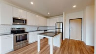 This single-floor unit showcases a thoughtfully designed, modern interior throughout. The kitchen serves as a central highlight, featuring crisp white cabinetry extending to the ceiling, a white subway tile backsplash, and dark granite countertops. A central island with matching granite provides additional workspace and casual dining potential. Stainless steel appliances, including a range, microwave, refrigerator, and dishwasher, complete the well-equipped space. Wood-look luxury vinyl flooring flows through the kitchen and living areas, adding warmth and continuity.

The living area is bright and open, benefiting from a large sliding glass door that leads to a private balcony overlooking an urban streetscape, allowing ample natural light to fill the space. The bedroom offers plush neutral carpeting, a large window with city views, and access to a spacious walk-in closet outfitted with wire shelving for organized storage.

The bathroom is cleanly appointed with a white vanity offering generous cabinet storage, a large mirror with modern strip lighting above, and a full soaking tub with shower. The consistent use of light tones, wood-look flooring, and recessed lighting throughout creates a cohesive, contemporary ambiance.