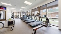 This fitness center in Wichita, Kansas, offers a comprehensive, single-floor layout designed to accommodate a wide range of workout needs. The space is finished with durable speckled rubberized flooring throughout, providing both safety and comfort underfoot. A drop panel ceiling with recessed fluorescent lighting ensures the entire area is evenly and brightly lit.

The main workout area is generously sized and lined along one side with large windows fitted with roller shades, allowing natural light to filter in while offering views of the surrounding outdoor landscape. A row of modern cardio equipment—including treadmills, ellipticals, and a rowing machine—runs along the windowed wall, making workouts visually engaging.

On the opposite side, full-length mirrors visually expand the space and support strength training. A cable and pulley multi-functional rack, adjustable bench, dumbbell rack, and a plyo box provide versatile options for resistance and functional training. A colorful array of medicine balls and additional free weights are also available.

Near the entry, a wall-mounted water bottle filling station and drinking fountain offer convenient hydration access. Mounted flat-screen televisions near the entrance area add entertainment. A door connects to an adjacent restroom facility, completing the practical amenities of this thoughtfully designed fitness space.