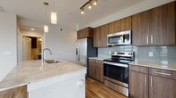 This contemporary single-floor residence is designed with a clean, modern aesthetic and thoughtful attention to detail throughout. The open-concept kitchen serves as a focal point, featuring rich walnut-toned cabinetry with sleek metal hardware, light granite countertops, a glass tile backsplash, and a full suite of stainless steel appliances including a French door refrigerator, electric range, and over-the-range microwave. A generous kitchen island with an integrated sink and pendant lighting above adds both functionality and style. Wood-style flooring extends through the main living and dining areas, where large windows dressed with horizontal blinds allow ample natural light to fill the space. A ceiling fan adds comfort to the spacious living area. The bedrooms offer a quieter retreat, finished with soft carpeting and windows that provide views of the surrounding area. The primary bathroom is well-appointed with a dual-sink vanity topped in granite, dark wood cabinetry, a large framed mirror, and a tiled walk-in shower. The neutral color palette and consistent finishes throughout give the residence a cohesive and polished character, making it a comfortable and stylish living environment.