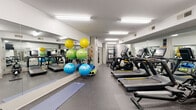 This fitness center is thoughtfully designed to accommodate a wide range of workout styles within a single, elongated space. The layout is organized to maximize functionality, with distinct zones dedicated to cardio, strength training, and functional fitness. Durable rubber flooring runs throughout, providing traction and shock absorption across all areas.

The cardio zone lines one side of the room and features a row of Matrix treadmills and elliptical machines, each equipped with individual screens for entertainment during workouts. Floor-to-ceiling mirrors run along the walls, visually expanding the space and allowing users to monitor their form. Overhead, flush-mounted fluorescent lighting ensures consistent, bright illumination throughout.

The strength and functional training area includes a multi-tiered dumbbell rack stocked with a wide range of weights, a cable and suspension training system, and an adjustable workout bench. Spin bikes with digital displays provide an additional cardio option, positioned near a wall-mounted flat-screen television.

A dedicated stretching and flexibility zone features yoga mats and a colorful collection of stability balls in multiple sizes, neatly organized on a rack. A motivational typographic mural featuring fitness-related words serves as a focal point, adding visual energy to the space. The overall ambiance is clean, modern, and purpose-driven.