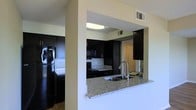 This single-floor space showcases a well-appointed kitchen designed for both functionality and style. The kitchen is outfitted with rich, dark espresso-toned cabinetry that extends from floor to ceiling, providing ample storage throughout. Granite countertops in a speckled light grey tone add a polished, durable work surface, while a matching granite breakfast bar with an undermount sink and modern stainless steel faucet creates a natural divide between the kitchen and the adjacent living area. This pass-through design enhances the open feel of the space, promoting easy interaction between rooms. Black appliances, including a full-size refrigerator, electric range, and built-in microwave, complement the dark cabinetry for a cohesive aesthetic. A tile backsplash adds a subtle textural element behind the range. The ceiling features a textured finish, and HVAC vents are positioned for efficient climate control throughout. Overhead lighting keeps the kitchen well-lit. A louvered door visible in the background hints at additional storage or utility space nearby. The overall ambiance is modern and sleek, with a strong contrast between the dark cabinetry and lighter countertop and wall tones.