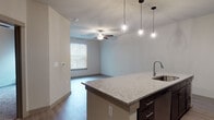 This single-floor residence offers a thoughtfully designed open-concept layout that seamlessly connects the kitchen and living areas. The kitchen serves as the centerpiece, showcasing an expansive granite-topped island with an undermount sink, complemented by rich dark espresso cabinetry extending to the ceiling and a classic white subway tile backsplash. Stainless steel appliances — including a refrigerator, range, and over-the-range microwave — complete the space. Geometric pendant lights hang above the island, adding a contemporary design touch. Wood-look flooring runs throughout the main living areas, while the living room benefits from natural light through a window and is finished with a ceiling fan for comfort. The bedrooms feature plush wall-to-wall carpeting and neutral wall tones, creating a calm and restful atmosphere. A generously sized walk-in closet with multiple shelving tiers and hanging rods provides substantial storage. The bathroom is finished with a granite-topped dark vanity, a framed mirror, and tile flooring, maintaining the consistent design language found throughout the unit. The overall aesthetic is clean, modern, and move-in ready.