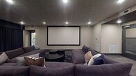 Located at 265 E State St in Columbus, Ohio, this single-floor screening and entertainment lounge offers a well-appointed, cinema-inspired environment designed for both relaxed viewing and social gatherings. The centerpiece of the room is a large wall-mounted projection screen paired with a ceiling-mounted projector, delivering an immersive viewing experience. A generously sized, deep-cushioned sectional sofa in charcoal gray anchors the seating area, complemented by a low round coffee table and layered area rugs that define the space. Recessed lighting throughout the drop tile ceiling provides controlled, dimmable illumination suited to screening events. Along the perimeter, bistro-style tables paired with orange upholstered chairs offer additional seating for dining or casual conversation, while a counter-height bar area with cabinetry and a compact refrigerator adds functional convenience. Floor-to-ceiling navy drapes along one wall enhance the room's cinematic atmosphere by managing light and adding a dramatic design element. Framed art pieces are displayed on the walls, adding a curated, gallery-like quality to the space. A glass-paneled entry door and smart-home control panel complete the modern, polished aesthetic of this versatile entertainment venue.