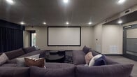 Located at 265 E State St in Columbus, Ohio, this single-floor screening and entertainment lounge offers a well-appointed, cinema-inspired environment designed for both relaxed viewing and social gatherings. The centerpiece of the room is a large wall-mounted projection screen paired with a ceiling-mounted projector, delivering an immersive viewing experience. A generously sized, deep-cushioned sectional sofa in charcoal gray anchors the seating area, complemented by a low round coffee table and layered area rugs that define the space. Recessed lighting throughout the drop tile ceiling provides controlled, dimmable illumination suited to screening events. Along the perimeter, bistro-style tables paired with orange upholstered chairs offer additional seating for dining or casual conversation, while a counter-height bar area with cabinetry and a compact refrigerator adds functional convenience. Floor-to-ceiling navy drapes along one wall enhance the room's cinematic atmosphere by managing light and adding a dramatic design element. Framed art pieces are displayed on the walls, adding a curated, gallery-like quality to the space. A glass-paneled entry door and smart-home control panel complete the modern, polished aesthetic of this versatile entertainment venue.