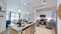 This thoughtfully designed one-bedroom, one-bathroom residence features a seamless open-concept layout that connects the kitchen, dining, and living areas into a cohesive, light-filled space. Light wood-style flooring flows throughout, complementing the soft, neutral wall tones. The kitchen is appointed with two-tone grey and white cabinetry, granite countertops, a large center island with pendant lighting, subway tile backsplash, and a full suite of stainless steel appliances including a side-by-side refrigerator, range, microwave, and dishwasher. Track lighting overhead ensures the space is well lit. The dining area accommodates a round wood table and sits adjacent to the living room, which benefits from large windows and a glass door that flood the area with natural light. The bedroom offers a comfortable, well-proportioned retreat with soft carpet and a muted color palette. A dedicated walk-in closet provides ample storage with wire shelving systems lining the walls. The bathroom features a granite-topped vanity with dark cabinetry, a large mirror, and a full tub-shower combination with marble-style tile surround and wood-look flooring, rounding out this efficiently designed and modern residence.