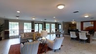 This single-floor space is thoughtfully designed to accommodate multiple functional zones within an open, connected layout. The living area serves as a central focal point, featuring a neutral-toned sofa accented with patterned throw pillows and a pair of stylish upholstered armchairs arranged around a glass-top coffee table. A warm-toned area rug anchors the seating arrangement, adding texture and contrast to the light tile flooring that extends throughout the space.

Natural light flows generously through a series of large windows dressed with curtains featuring subtle banding detail, offering views of greenery outside. Recessed lighting and flush-mount ceiling fixtures provide consistent illumination throughout. Crown molding along the upper walls adds a refined architectural touch.

Adjacent to the living area, a dining space accommodates a table with upholstered seating, maintaining the space's cohesive, understated palette. Further along, a dedicated bar or beverage area is visible, complete with shelving and counter seating, adding a social and functional dimension to the layout. The overall ambiance is polished and welcoming, blending comfort with a clean, contemporary sensibility.