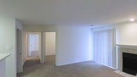 This single-story space presents a straightforward, functional layout with a calm, neutral aesthetic throughout. The main living area is finished with wall-to-wall beige carpeting, which lends a soft, understated feel underfoot. Walls are painted in a clean, off-white tone that reflects light evenly across the room, contributing to a bright and airy atmosphere.

A large window fitted with vertical blinds allows generous natural light to filter into the space, illuminating the open living area. The blinds offer both privacy and light control, making the room adaptable to various lighting preferences throughout the day.

The ceiling is smooth and standard in height, with visible HVAC vents indicating a central air system. A hallway visible from the main living area leads to additional rooms, suggesting a practical, connected floor plan. The transition between spaces is defined by clean doorframes and consistent flooring, maintaining visual continuity throughout.

Overall, the space is clean, unfurnished, and move-in ready, offering a blank canvas that is well-suited for customization. The neutral finishes and carpeted floors create a comfortable, livable environment with a modest, functional character.