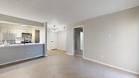 This efficiently designed one-bedroom, one-bathroom single-story residence offers a well-organized layout across its compact footprint. The open living room is a visual highlight, featuring distinctive herringbone-patterned luxury vinyl flooring in a light wood tone, neutral beige walls, and a textured ceiling. A tiled fireplace with a raised hearth anchors one wall of the living area, flanked by windows fitted with horizontal blinds that allow natural light to filter in. A modern ring pendant light illuminates the central hallway area.

The kitchen is positioned adjacent to the living space and is defined by a peninsula counter with a bright white surface and an undermount sink with a matte black faucet. Gray shaker-style cabinetry pairs with stainless steel appliances, including a refrigerator, gas range, and over-the-range microwave, all accented by track lighting overhead.

The bedroom features soft carpet flooring, a ceiling fan with light, and a large window providing natural light. The bathroom includes a single-sink vanity with gray cabinetry, white countertop, a framed mirror, overhead storage cabinet, and a full tub-shower combination. A closet and hallway complete the unit's practical layout.